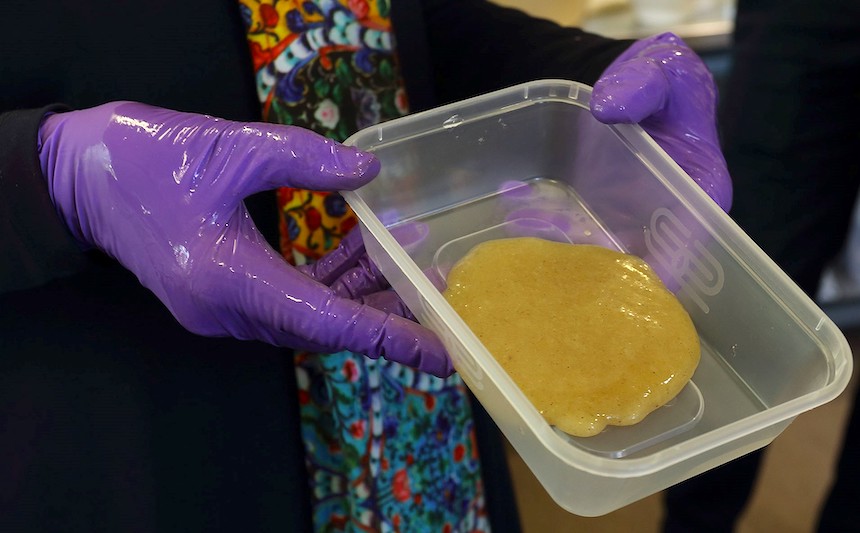 A person wearing plastic gloves displays a plastic tub containing a dark yellow substance