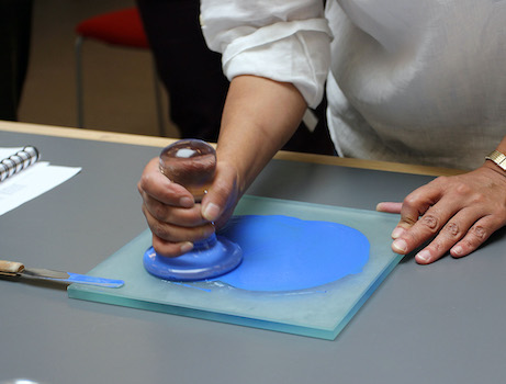 A hand mills bright blue powder into paint on a glass surface