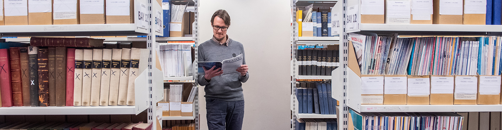 A person leans on a book shelf reading - there are full bookshelves either side of them