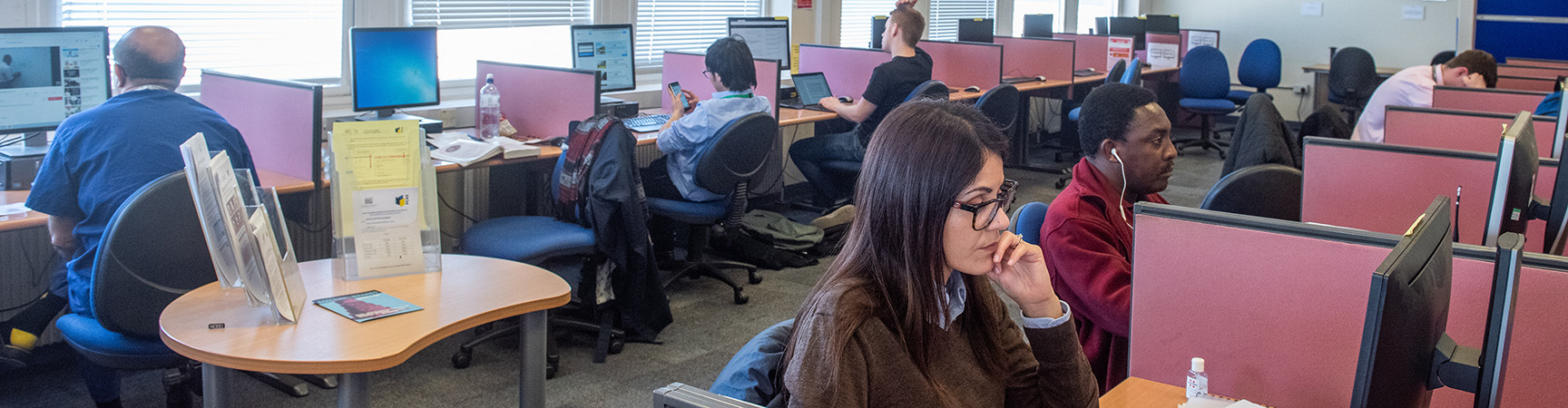 Students sit at individual desks each using a desktop computer, they are separated with red separators