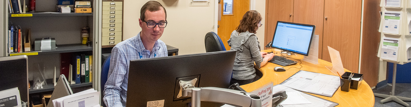 Two members of staff sit behind a curved wooden desk, each is looking at a desktop computer