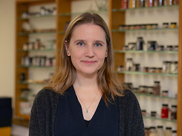 A woman with shoulder-length blonde hair wearing a dark top stands in front of wooden shelving