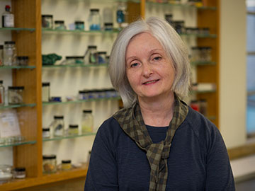 A woman with short grey hair wearing a dark jumper and checkered scarf sits in front of wooden shelving