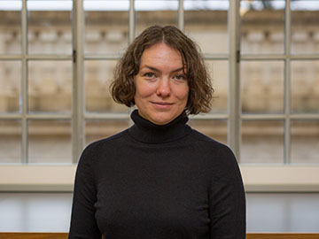 A woman with short hair wearing a black polo neck jumper stands in front of windows