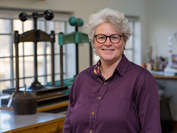A woman with short grey hair wearing a dark purple blouse stands next to a wooden table