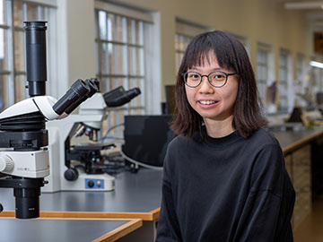 A woman with dark hair and glasses wearing a dark jumper sits next to a bench with microscopes