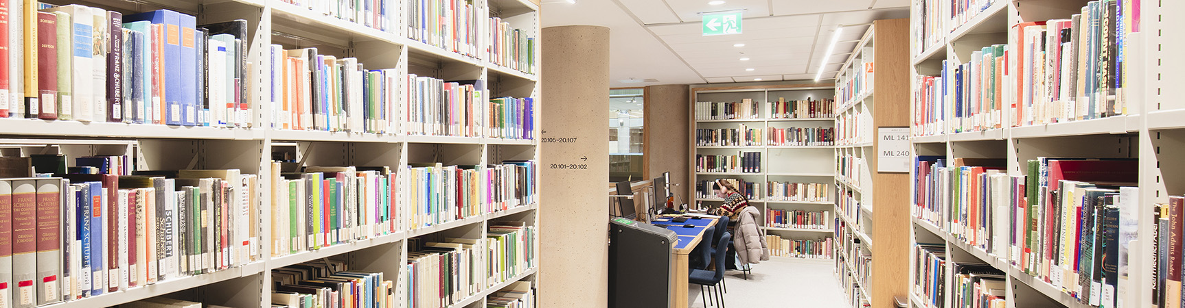 An aisle between full wooden bookshelves in the Humanities Library; at the end of the aisle is a soft seating area in which people are studying