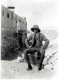 A black and white photograph of a man in a suit and hat sitting in front of a building