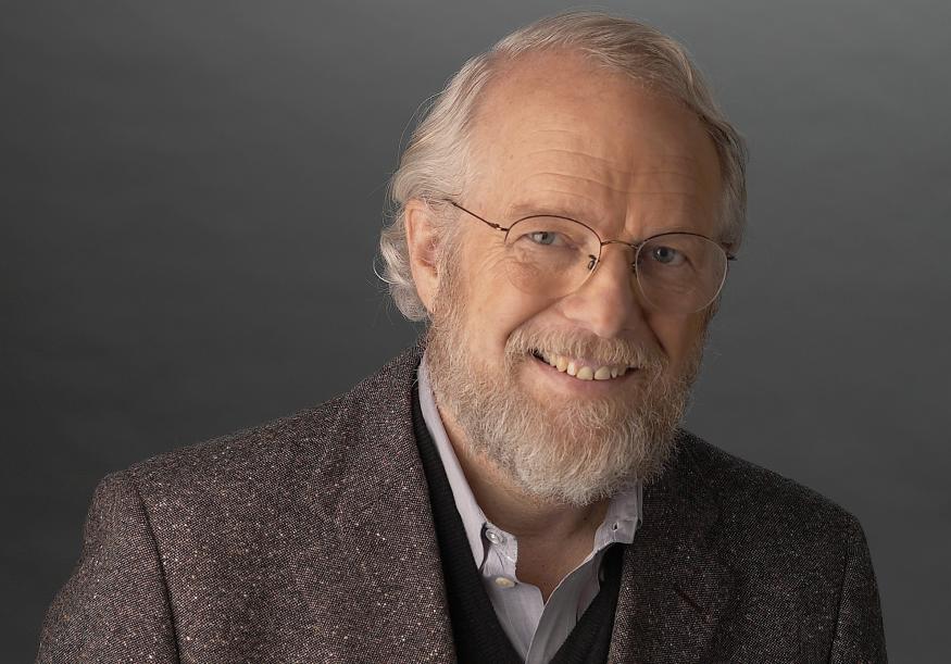 A smiling man with glasses wearing a pale purple shirt and dark brown jacket