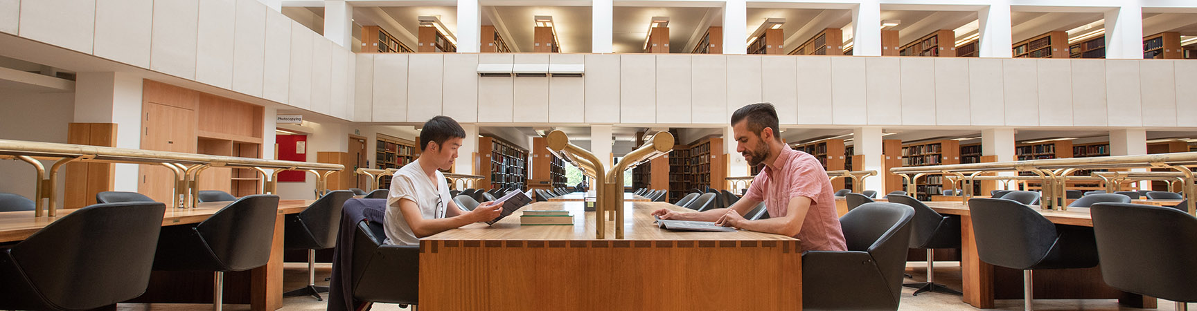 Two readers sit facing each other either side of a wooden desk, the room behind is light and airy