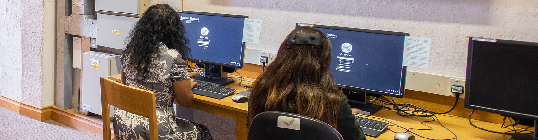 Two students sit next to each other each looking at a desktop computer screen