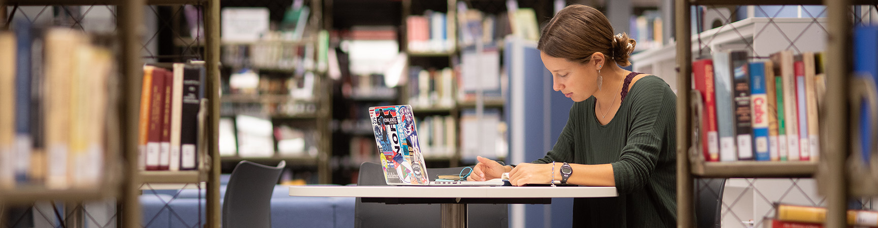 A student sits at a small white desk, surrounded by metal bookshelves