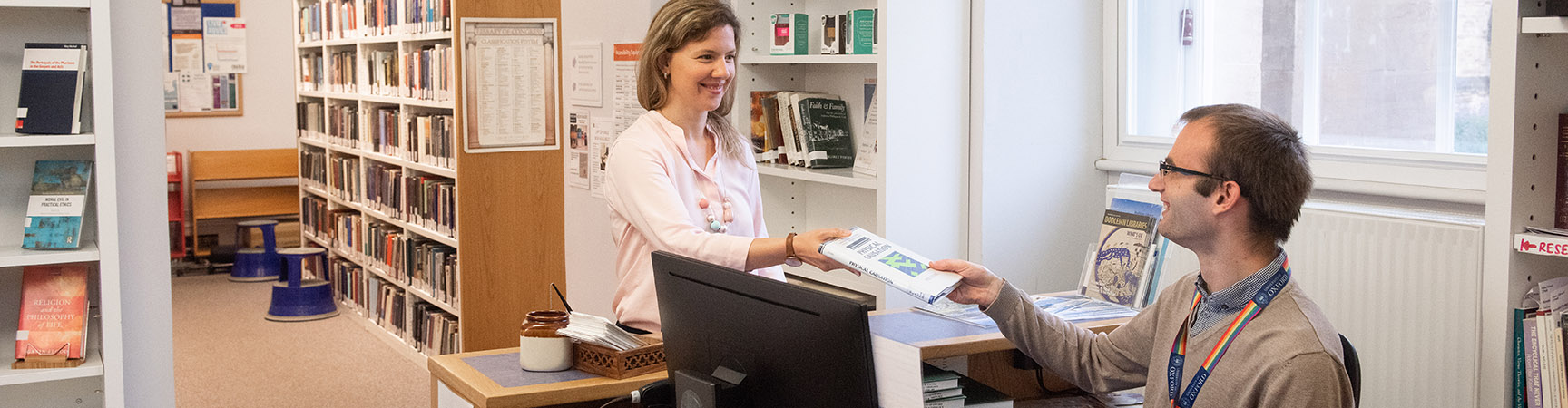 A librarian - sitting behind a computer at a desk - hands a book to a student standing behind the desk