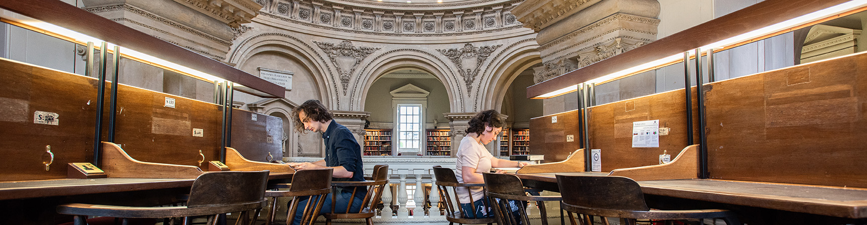 Two students sit back to back, they are each working on long wooden desks