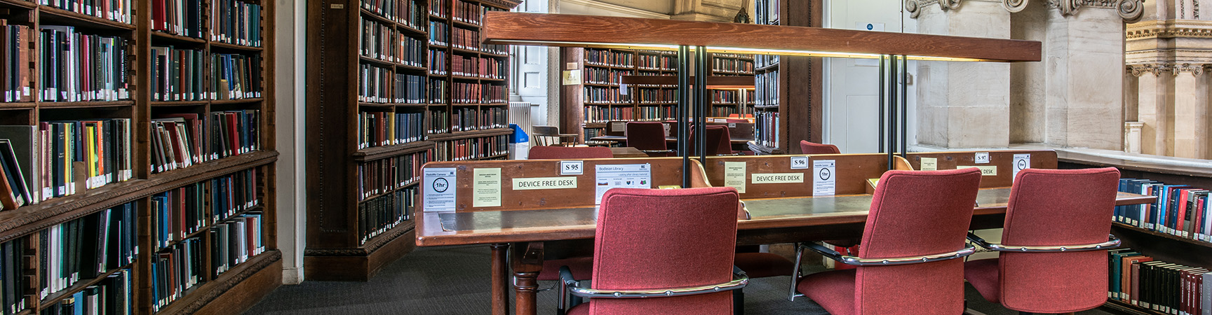 A row of three pale red chairs in front of a desk, bookshelves are in the background