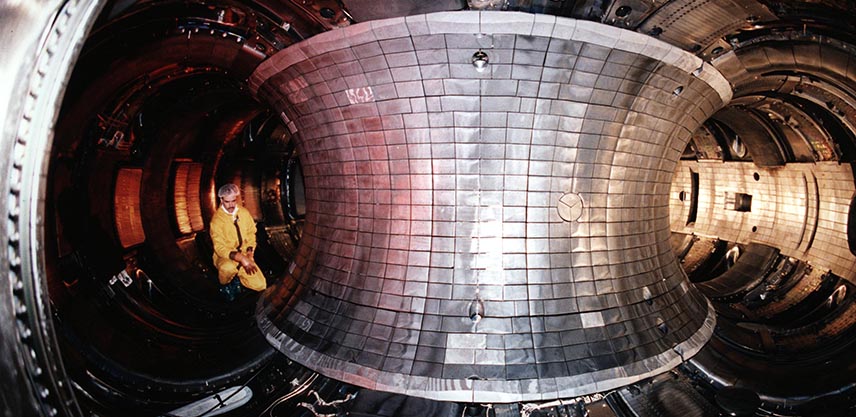 A view inside a giant silver metal doughnut. The floor is curved and black with some wires, the central wall of the ‘hole’ is silver tiles. On the left a man in a yellow jumpsuit kneels on the floor. The wall behind him has orange glowing squares.
