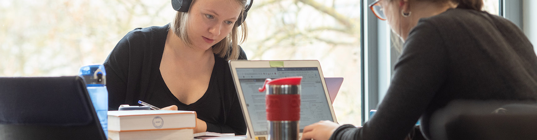 Two students work opposite each other at a desk. One has a KeepCup