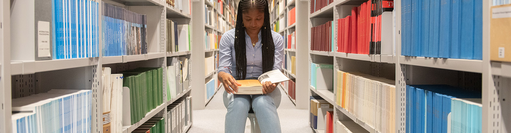 A woman sitting on a stool between bookshelves reading a book