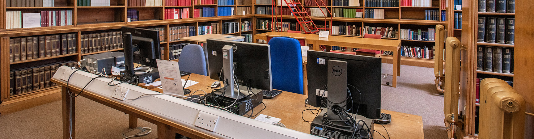 A row of desktop computer on a wooden desk with a bookshelf on the back wall
