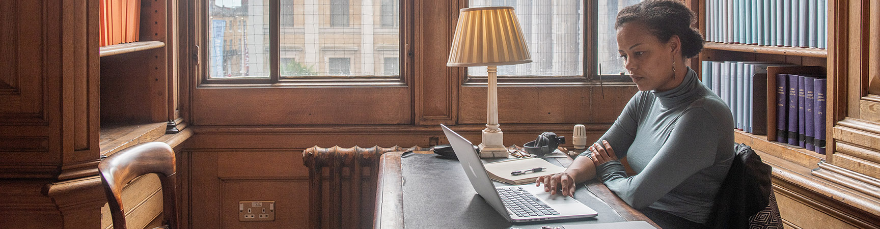 A student sit in a small alcove desk - there is a small lamp on the table and the student is working on a laptop