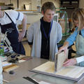 Two people look on as a conservator holds a manuscript upwards and affixes it to the cover laid out underneath it