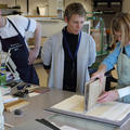 Two people look on as a conservator holds a manuscript upwards and affixes it to the cover laid out underneath it