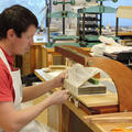 A man sits at a desk. In front of him is a wooden frame - a manuscript sits at the base. Threads run from the top of the frame to the bottom as the man sews between them