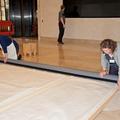 Two people kneel on the floor at two corners of the tapestry - they are gently rolling it up ready to be moved