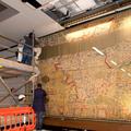 A floor to ceiling height glass case with a tapestry map behind it. A scaffold is in front of the case whilst the installation is completed