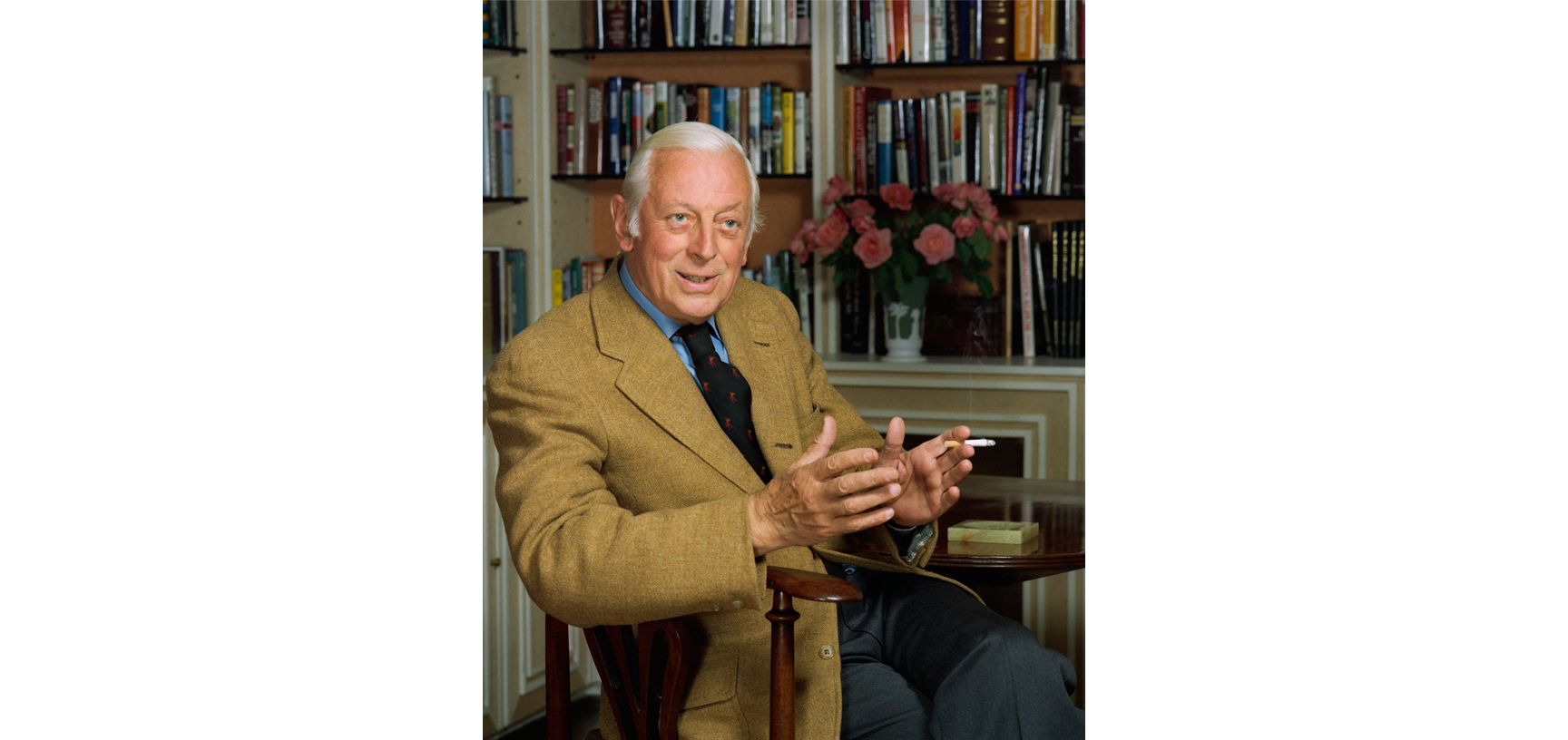Portrait of Alistair Cooke - an older white male with white hair, dressed in a tan blazer, blue shirt and navy tie, sitting in a wooden chair looking diagnol to the camera with bookshelves in the background