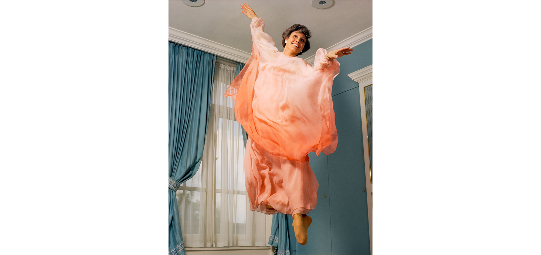 Portrait of Angela Rippon - a young white woman with short brown hair, dressed in a peach chiffon dress, jumping in the air, arms outstreched and toes pointed, looking into the distance