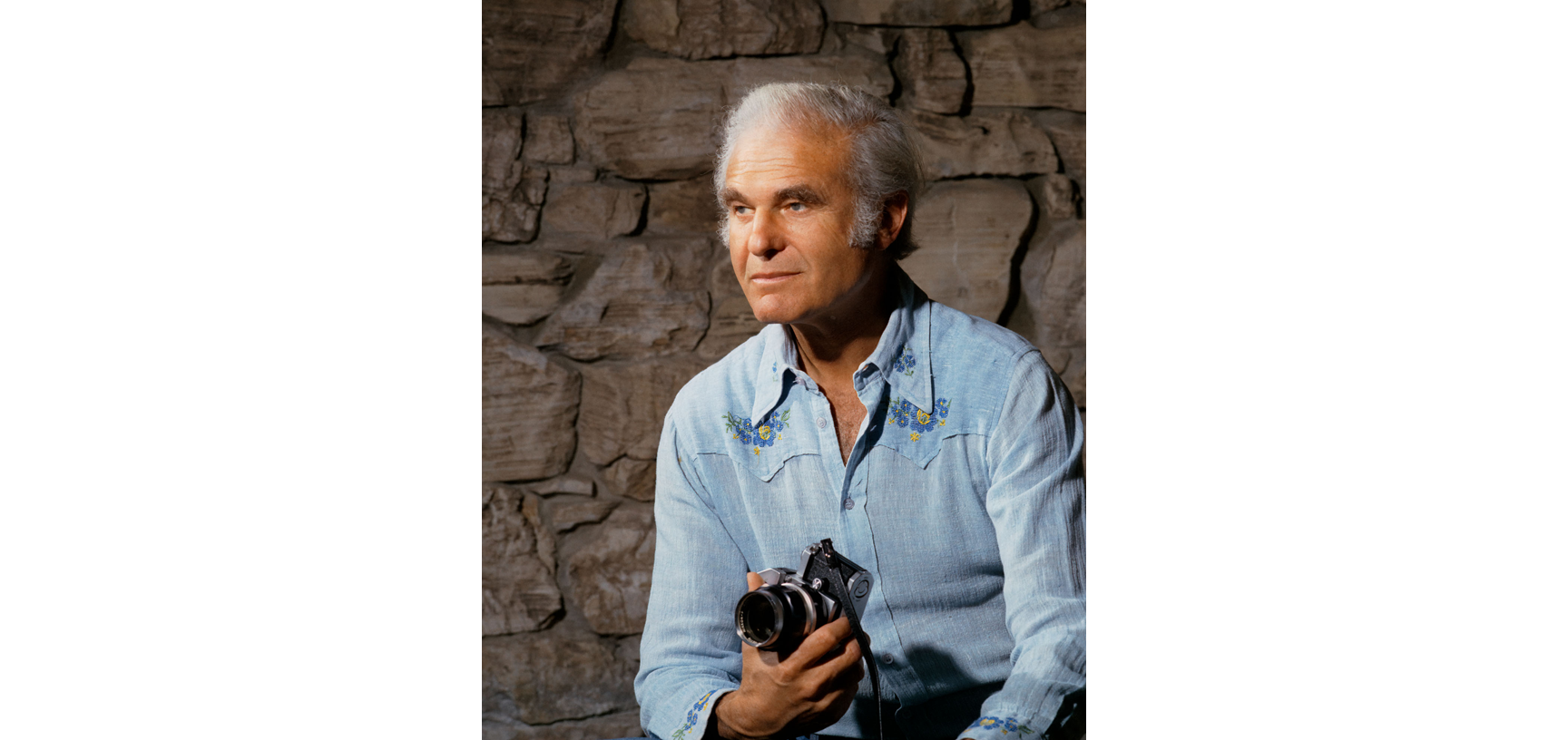 Portrait of Alan Lloyd Hodgkin - an older white male with white hair, dressed in a light blue shirt, sitting in front of a stone wall, holding a camera in one hand and looking off to the left