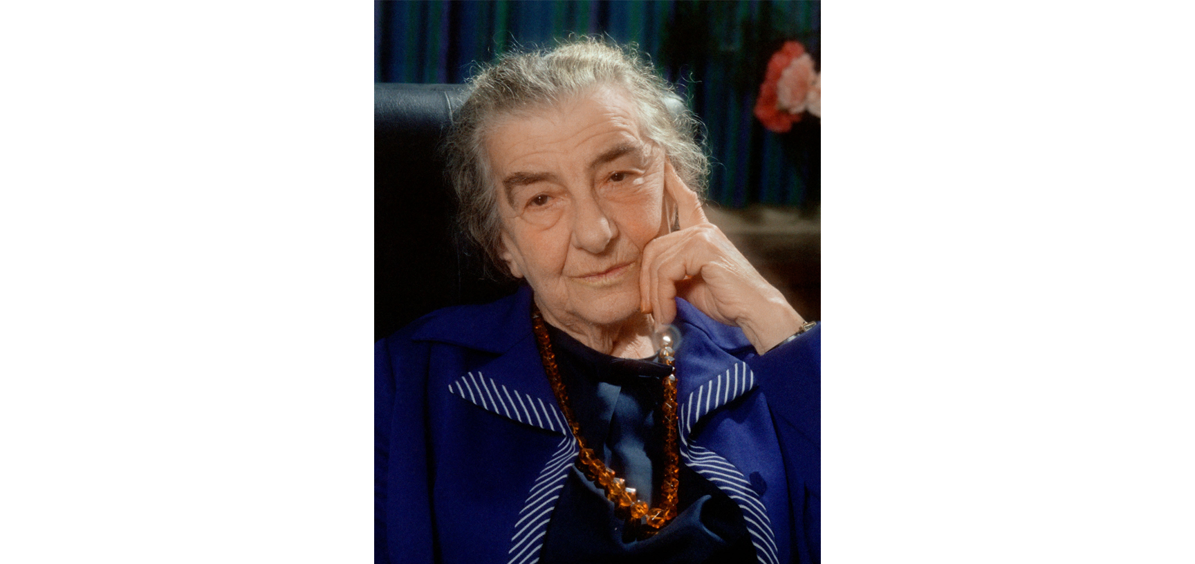 Portrait of Golda Meir - a older white woman with grey hair tied up, dressed in a navy dress and navy blazer with a brown beaded necklace, looking into the camera with her head resting on her hand