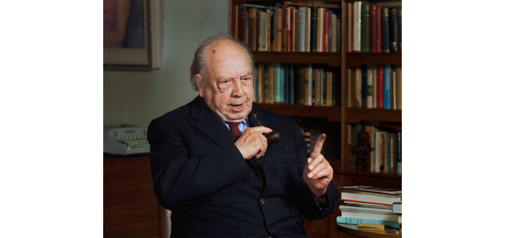 Portrait of JB Preistley - an older white male dressed in a navy blue suit, light blue shirt and red tie, sitting in a chair with bookshelves in the background diaganol to the camera
