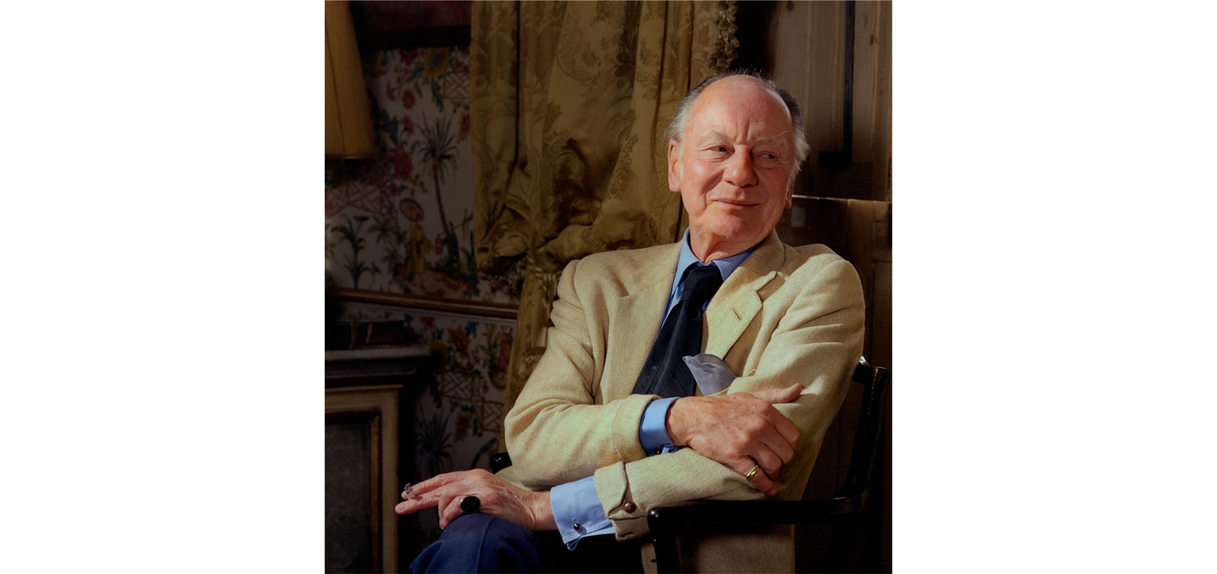Portrait of John Gielgud - an older white male balding with white hair, dressed in a cream blazer, blue shirt and navy tie, sitting in a wooden chair looking to the right with a tapestry in the background, holding a cigarette in one hand