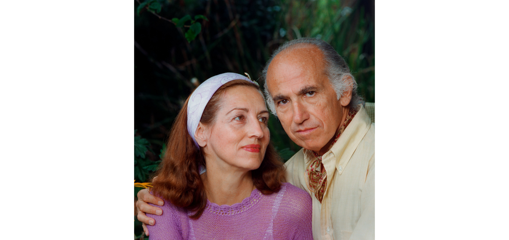 Portrait of Jonas Salk and Françoise Gilot - an older white male balding with white hair, dressed in a cream shirt and jumper with a patterned neckscarf, hs arm is round a young red haired woman in a pink jumper and white headband.