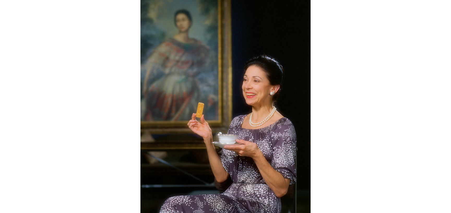 Portrait of Margot Fonteyn - a middle aged white woman with dark hair up in a bun, wearing a purple and white flower patterned dress, sitting in a chair with a cup of tea and biscuit in hand, sat in front of a large oil painting