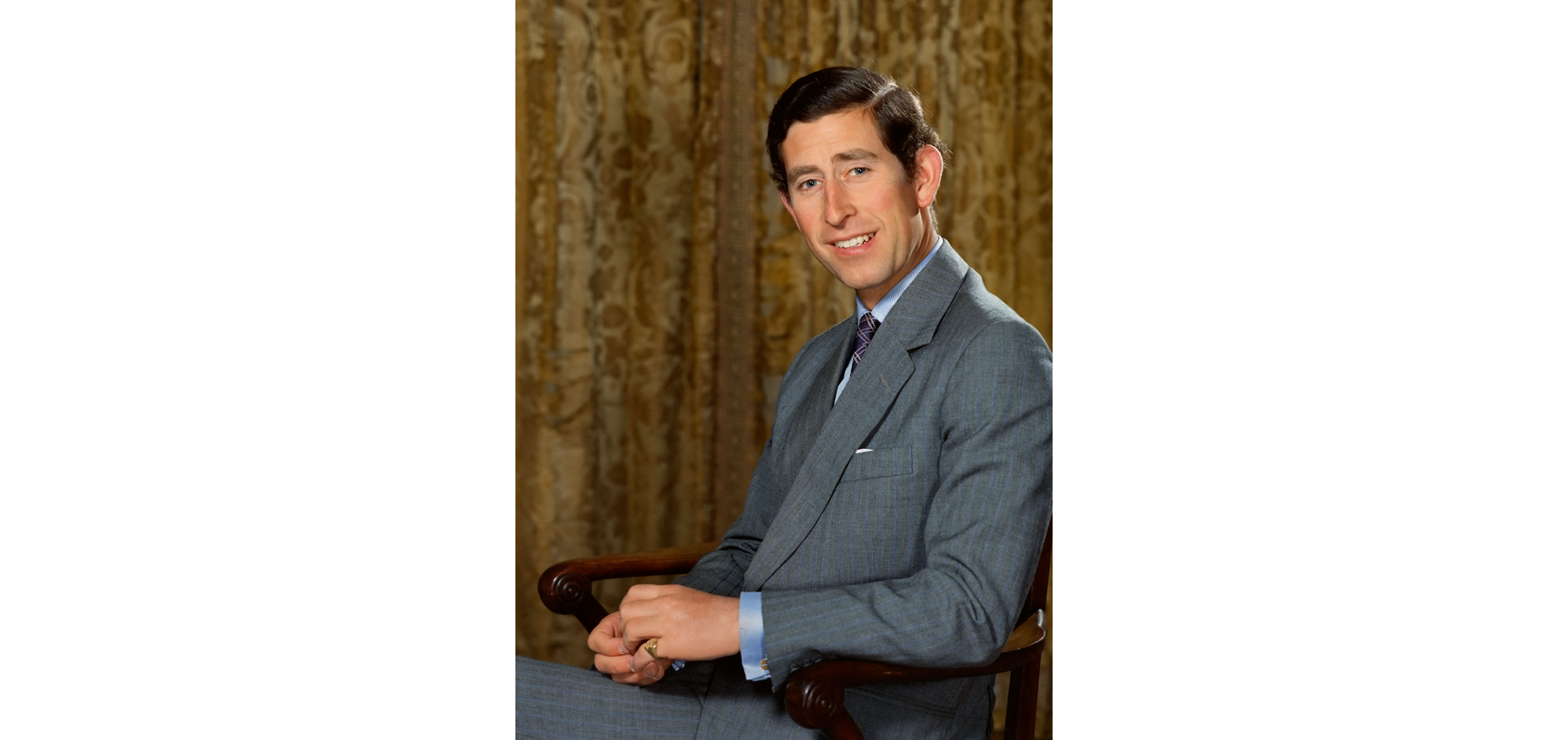 Portrait of Prince Charles - a young white male with dark hair and dressed in a grey suit, shirt & tie, sitting in a wooden chair looking straight at the camera