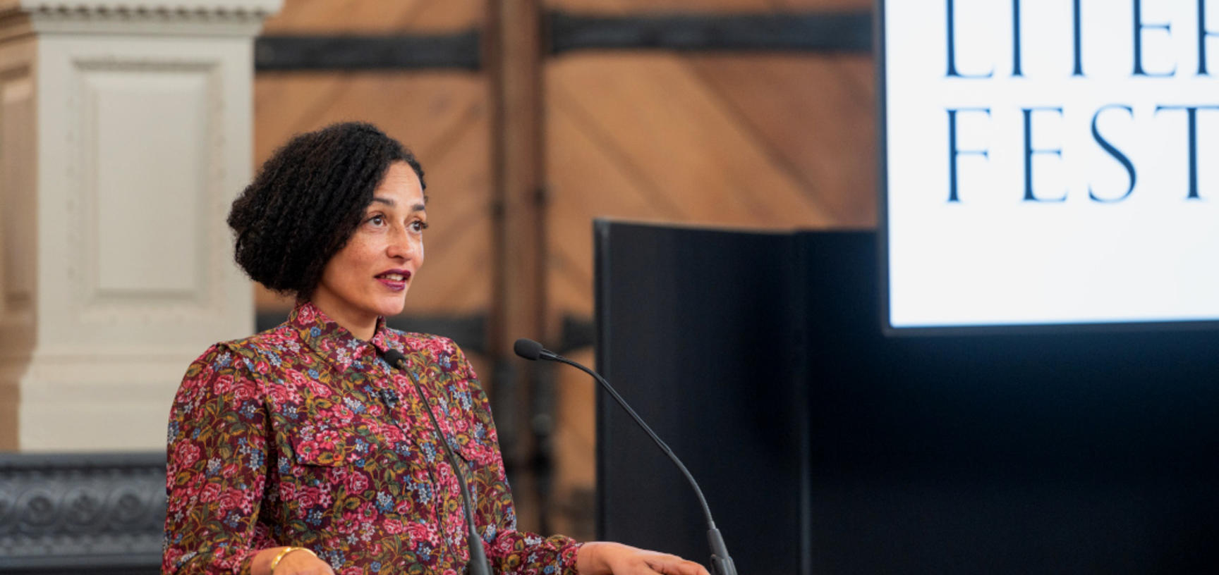 Zadie Smith speaks from a podium in front of a blue background, beside a screen