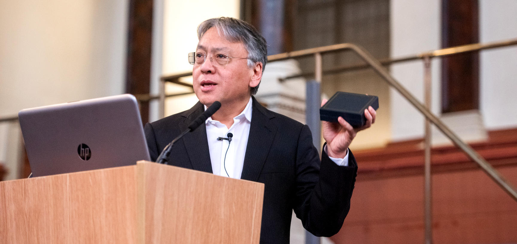 Sir Kazuo Ishiguro stands in front of a microphone and laptop, holding the Bodley Medal in its box
