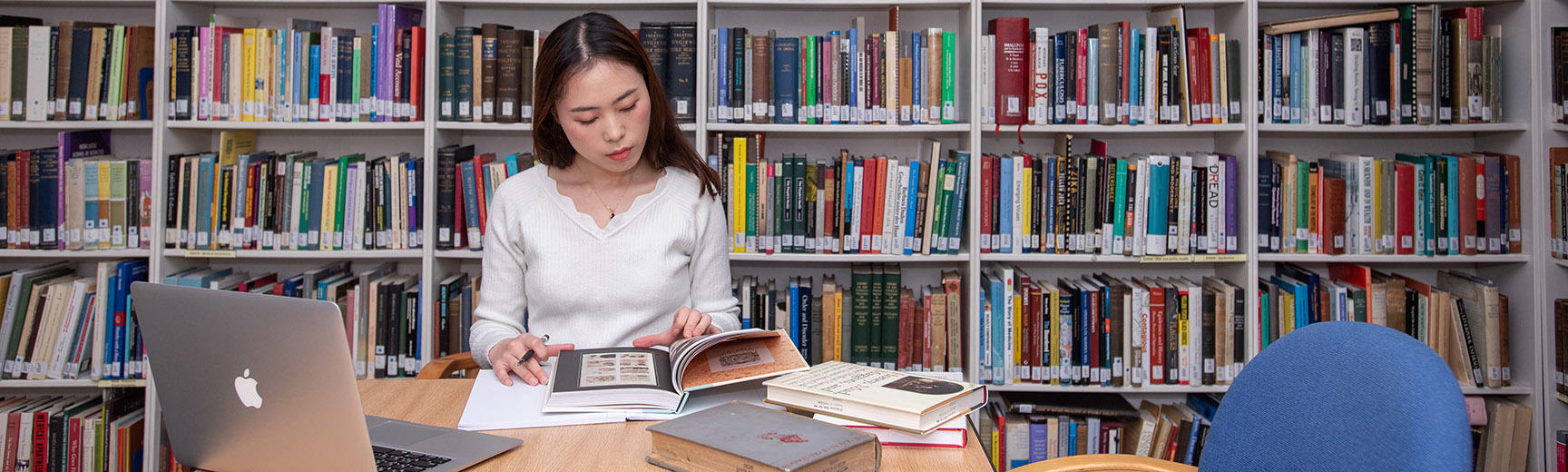 A student sits at a table - they look at a book and have an open laptop - behind is a bookshelf covering the whole wall