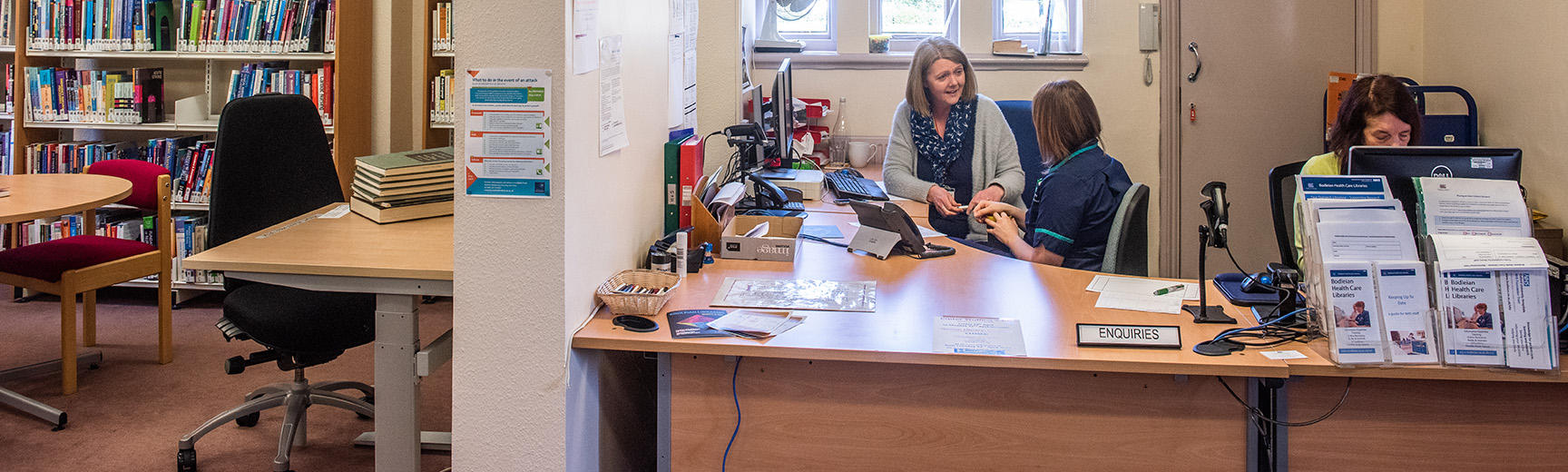 Two people sit behind a desk talking, one is dressed in an NHS nurse uniform. To the left the corner of the reading room can be seen with a table and bookshelves