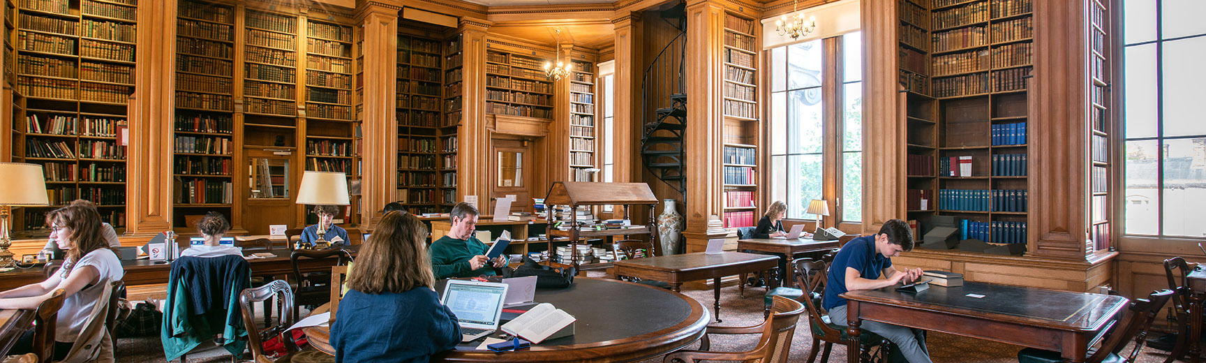 Students sit at individual desks in a wood-panelled room with bookshelves