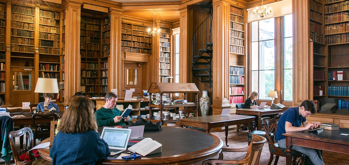 Students sit at individual desks in a wood-panelled room with bookshelves