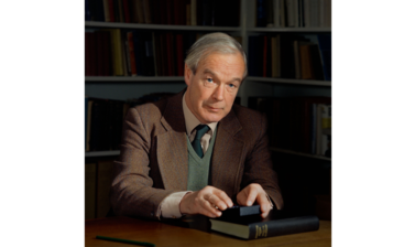 Portrait of Alan Lloyd Hodgkin - an older white male with white hair, dressed in a brown blazer, cream shirt, dark green navy tie and a green vest, sitting at a wooden desk looking at the camera with bookshelves in the background