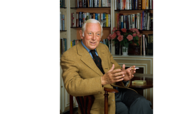 Portrait of Alistair Cooke - an older white male with white hair, dressed in a tan blazer, blue shirt and navy tie, sitting in a wooden chair looking diagnol to the camera with bookshelves in the background