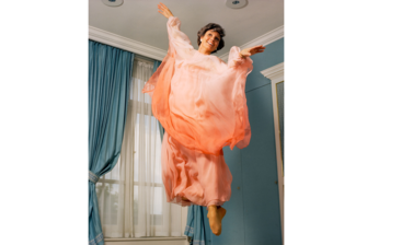 Portrait of Angela Rippon - a young white woman with short brown hair, dressed in a peach chiffon dress, jumping in the air, arms outstreched and toes pointed, looking into the distance