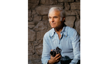 Portrait of Alan Lloyd Hodgkin - an older white male with white hair, dressed in a light blue shirt, sitting in front of a stone wall, holding a camera in one hand and looking off to the left