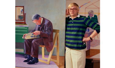 Portrait of David Hockney - a middle aged white man with blonde hair and glasses, wearing a stripey blue and green jumper and cream trousers, standing next to a large painting of an older man reading a newspaper, looking directly at the camera