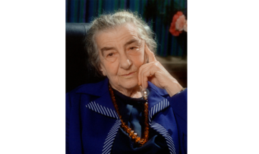 Portrait of Golda Meir - a older white woman with grey hair tied up, dressed in a navy dress and navy blazer with a brown beaded necklace, looking into the camera with her head resting on her hand